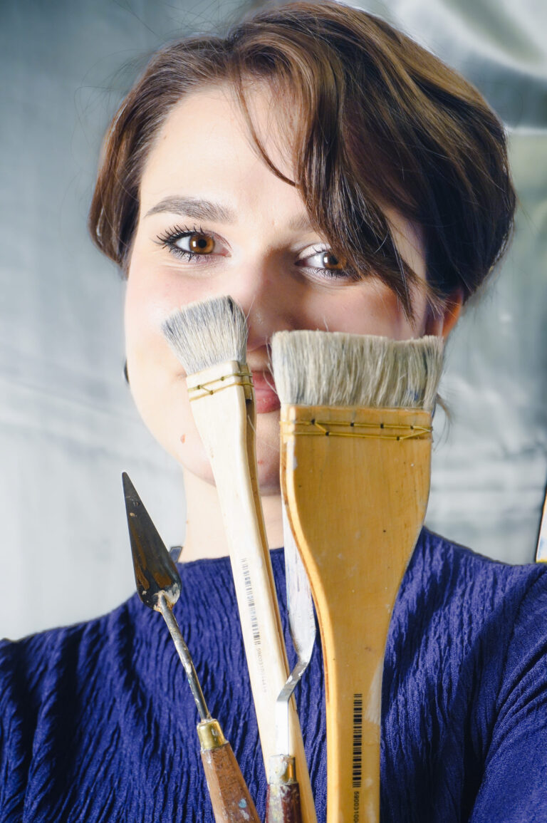 A female artist hiding behind her paintbrushes smiles at the camera.