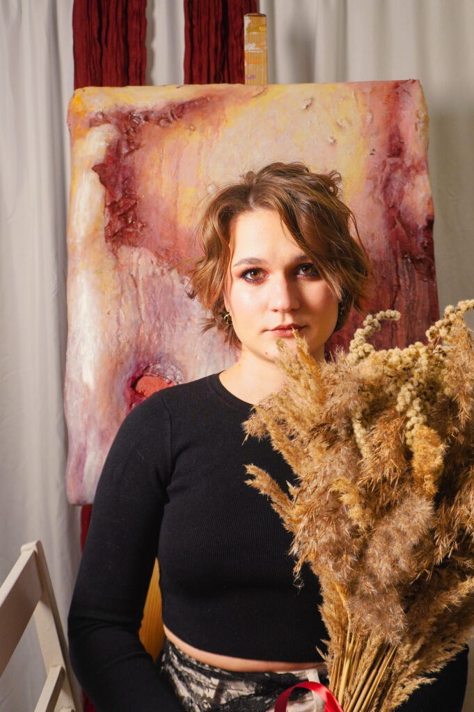 The artist sits with a bouquet of dried flowers in front of her painting.
